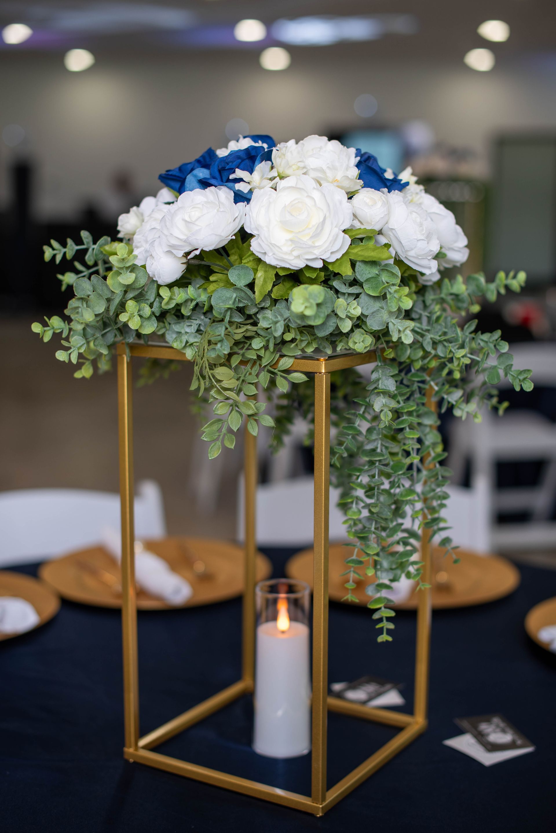 White floral centerpiece with greenery in a gold frame over a lit candle on a banquet table