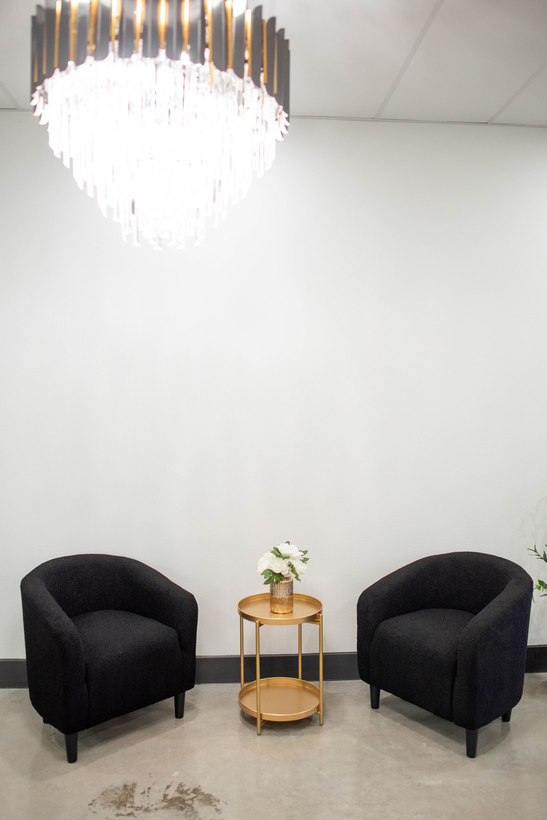 Modern lounge with two black chairs, a small round table, and a chandelier against a white wall.