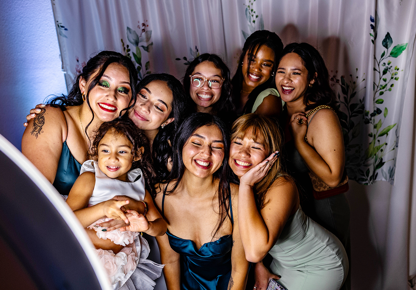 Group of eight people posing closely together indoors, smiling in colorful dresses before a curtain backdrop.