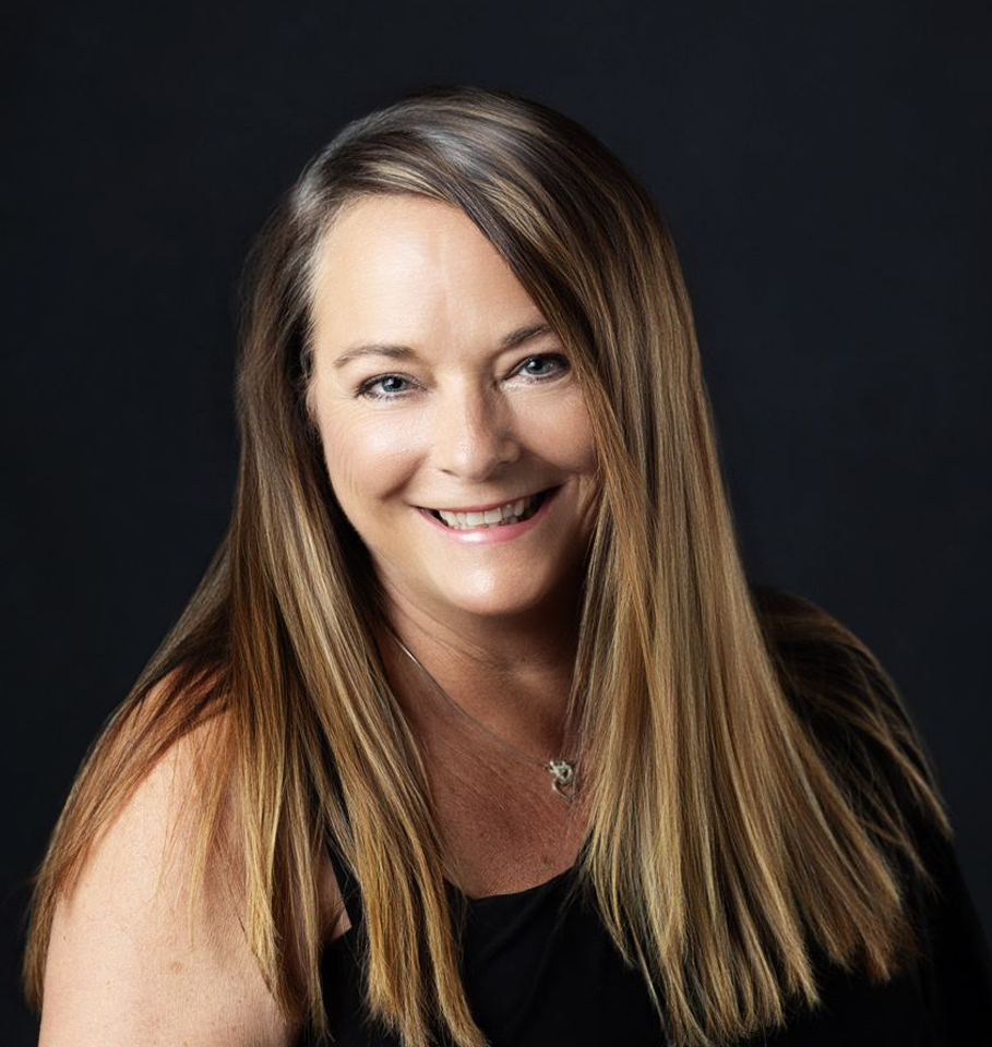 Woman with long brown hair smiles at the camera. Black top, dark background.