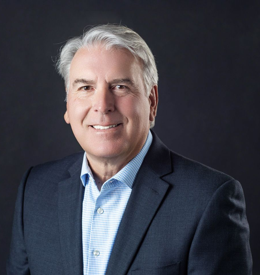 Mature man with gray hair, smiling, wearing a blue shirt and navy blazer against a dark background.