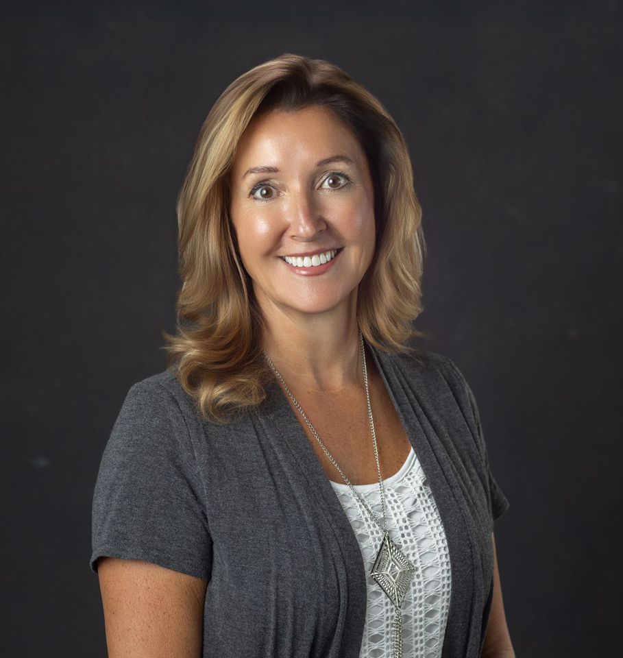 Woman with shoulder-length brown hair smiles, wearing a gray cardigan and white top, against a black background.