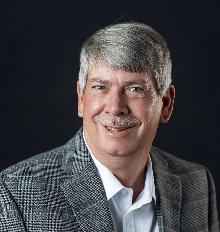 Middle-aged man with gray hair and mustache smiles, wearing a gray plaid blazer and white shirt against a dark background.