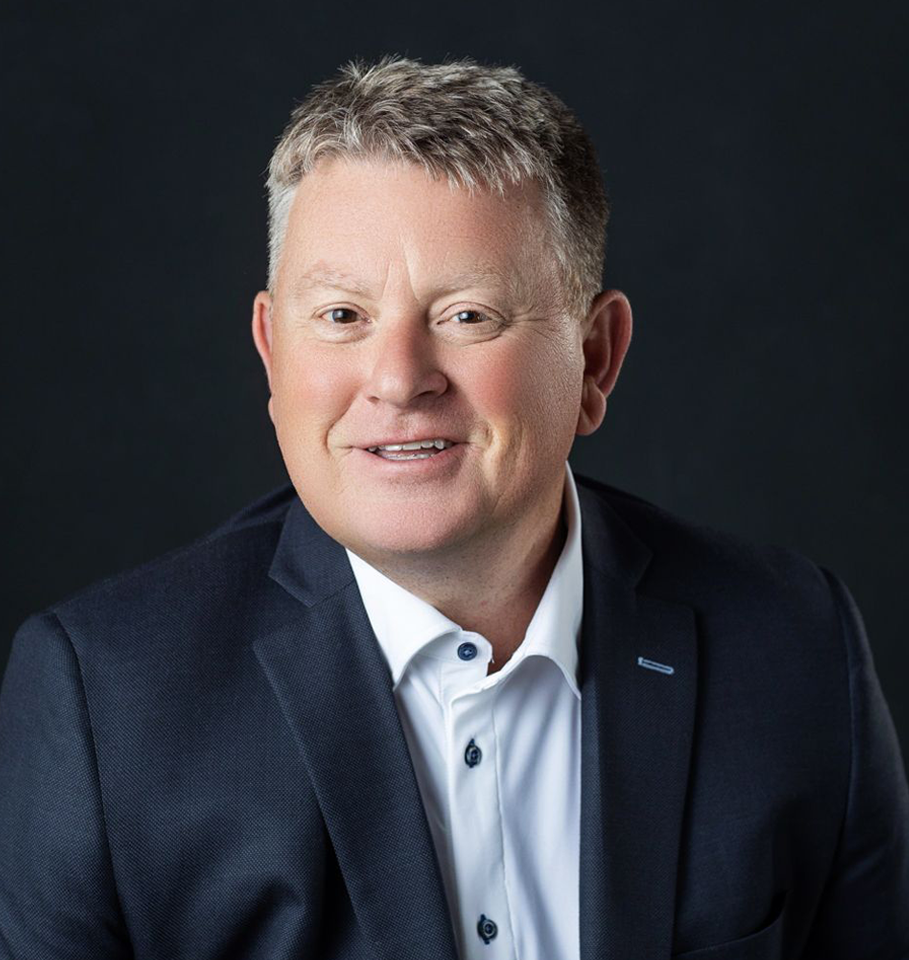 Man in a suit smiles at the camera against a dark background.