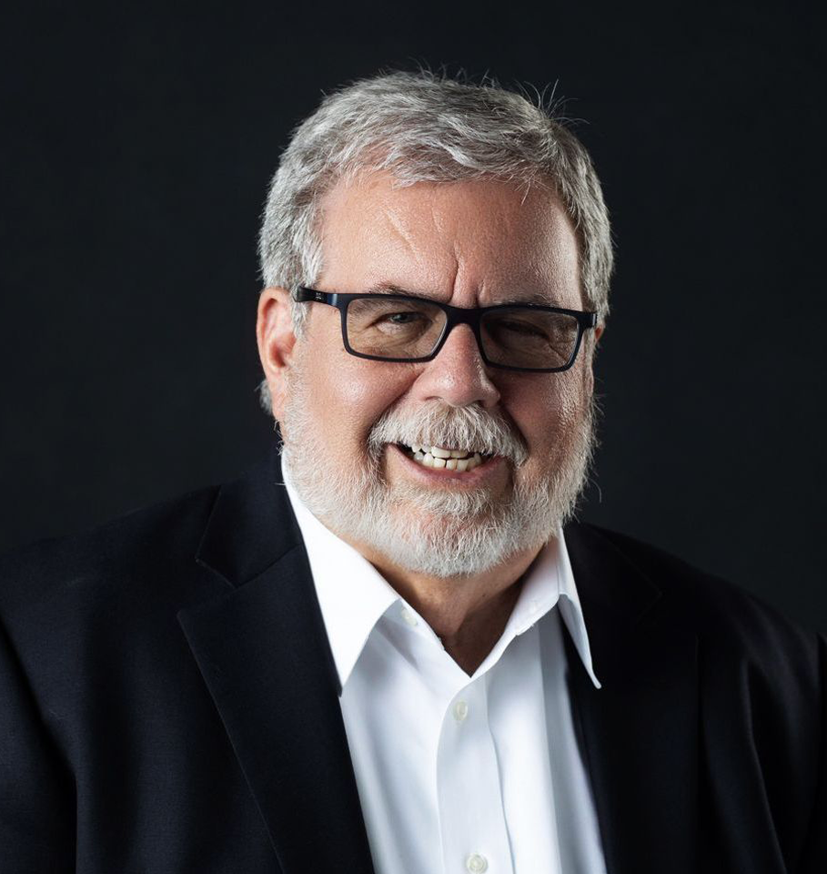 Bearded man with glasses smiling, wearing a dark suit and white shirt against a dark background.