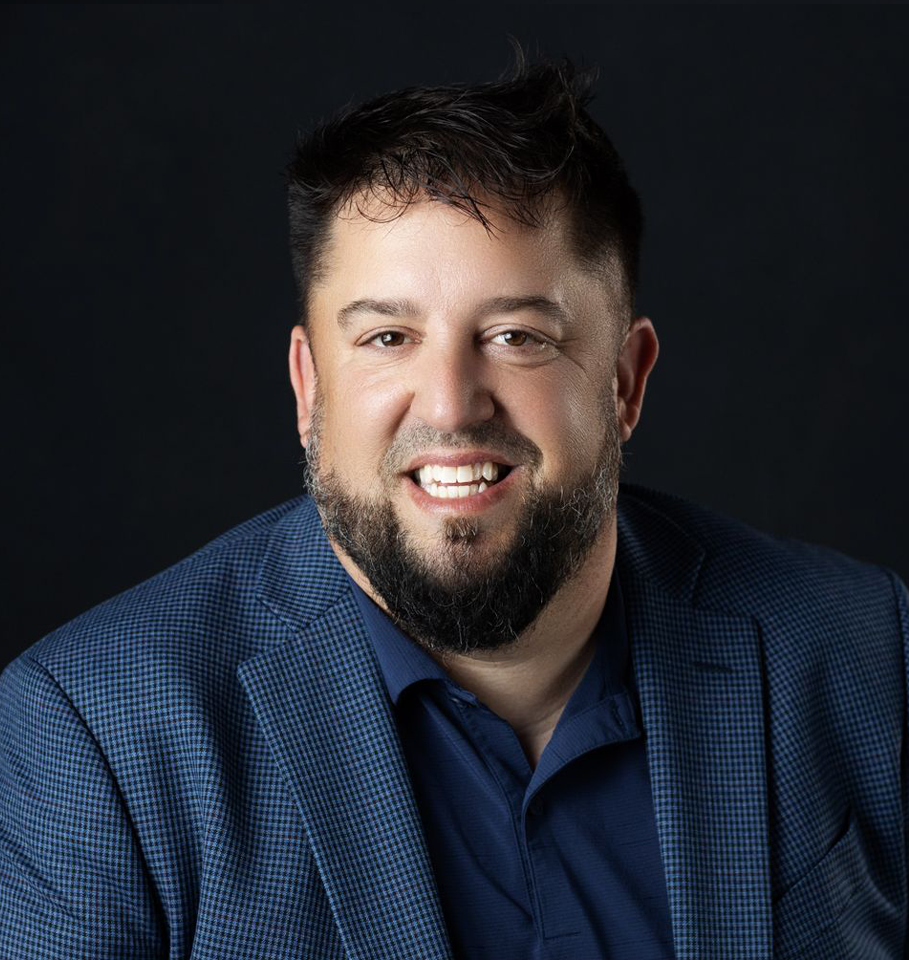 Smiling man with dark hair and beard, wearing a blue blazer and shirt, against a black backdrop.