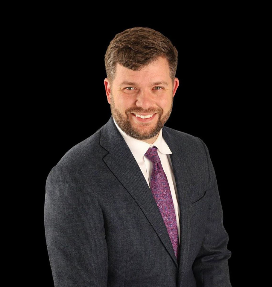 Man in a dark suit and purple tie smiles against a black background.