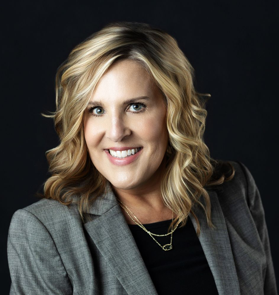 Smiling woman with blonde hair, in a gray blazer and black top against a dark background.
