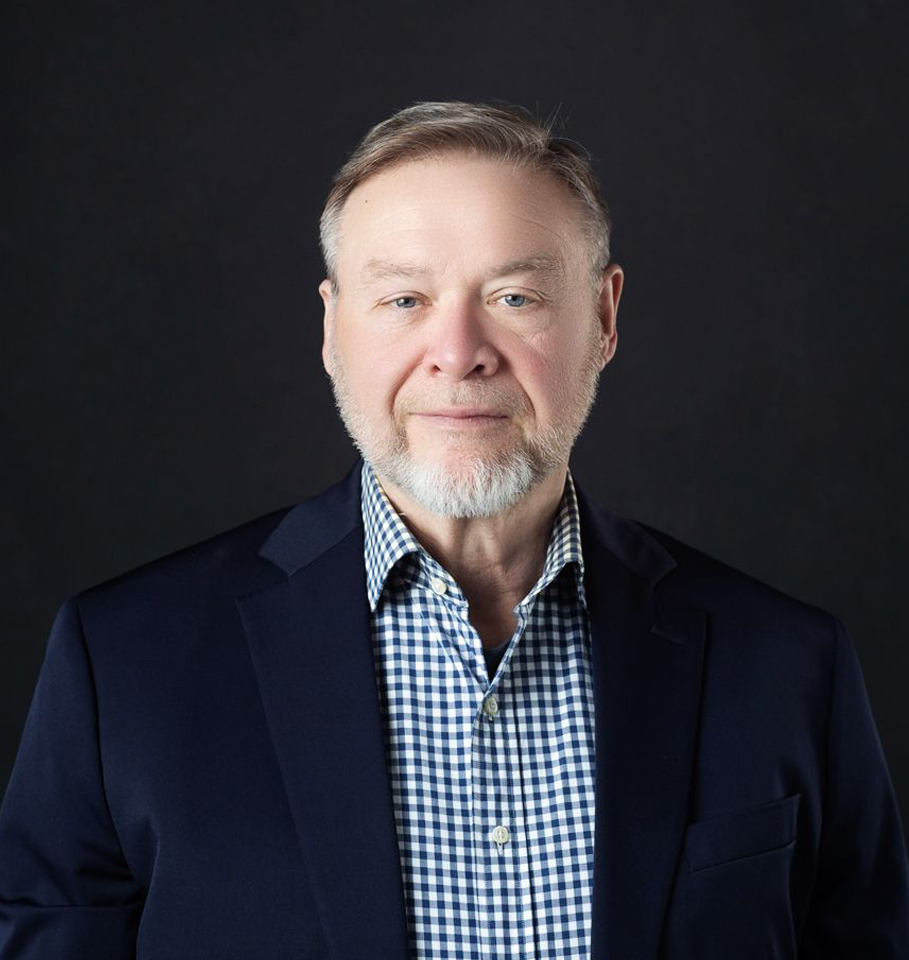A man with a gray beard and blue blazer smiles against a black background.
