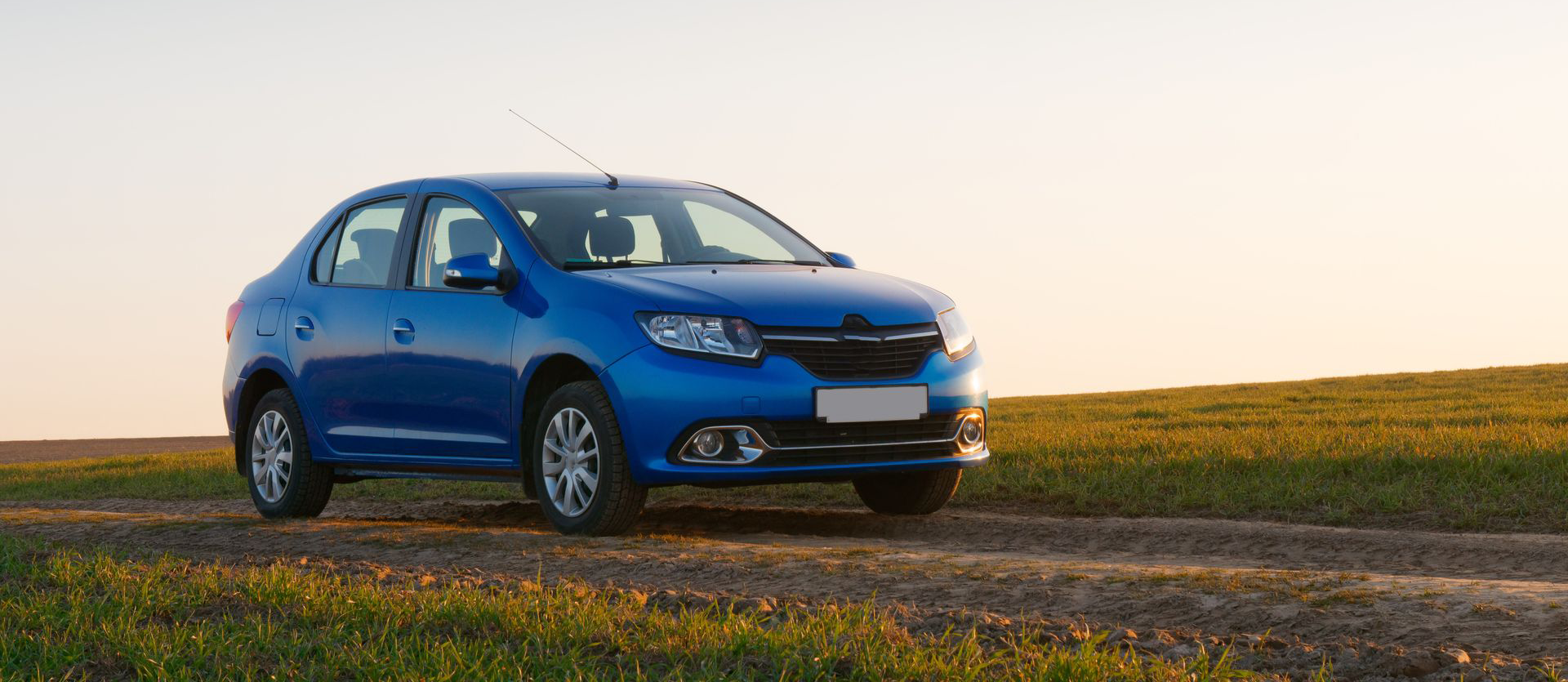 Blue car parked on a dirt road in a field with green grass under a clear sky.