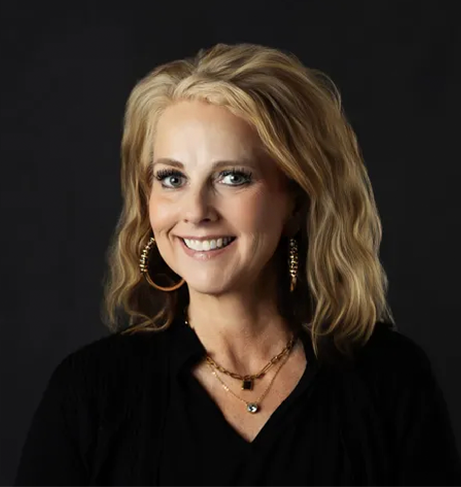 Woman with blonde hair smiles, wearing black top and gold jewelry against a dark background.