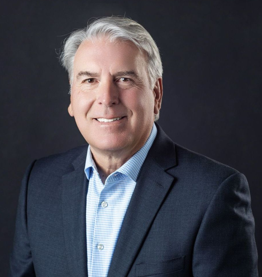 Man with gray hair, wearing a blue shirt and a navy blazer, smiling at the camera against a dark gray background.