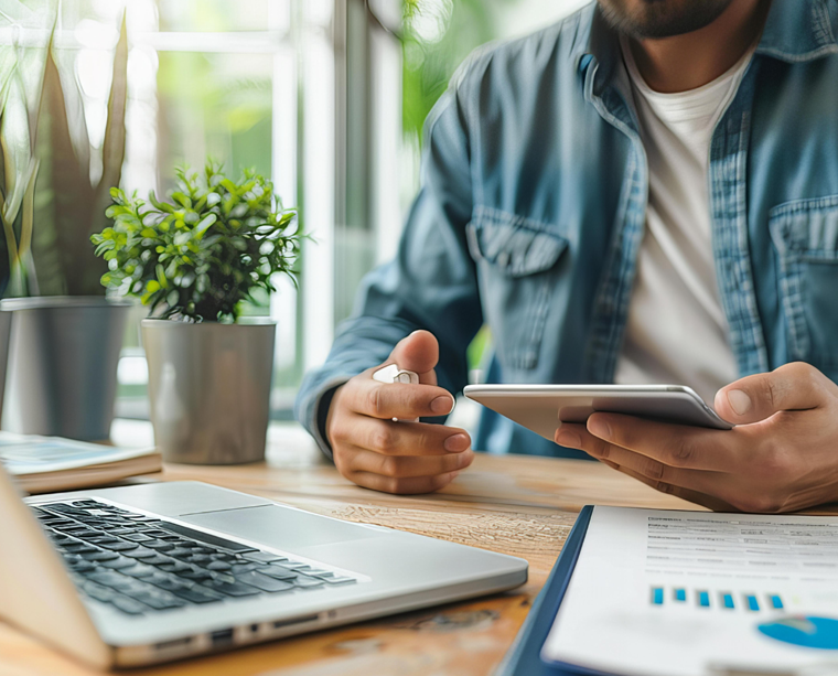 Man in denim shirt using a tablet at a desk with a laptop, potted plant, and documents.