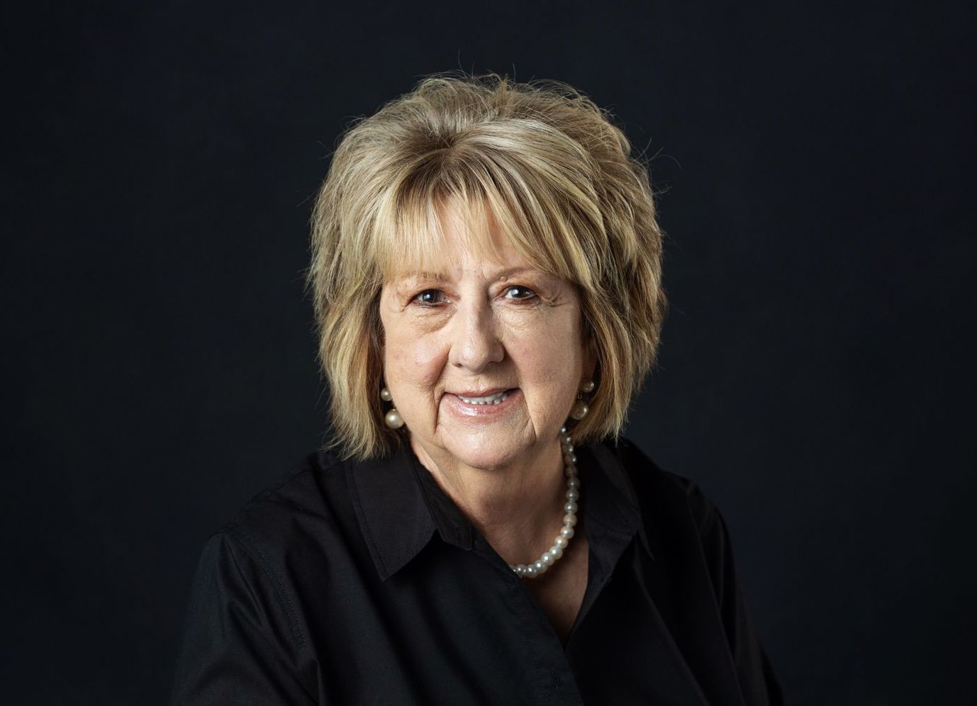 Woman with blonde hair and pearls, wearing a black shirt, smiles at the camera against a dark background.