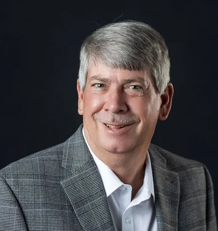 Man with graying hair, wearing a plaid blazer, smiles at the camera, against a dark background.