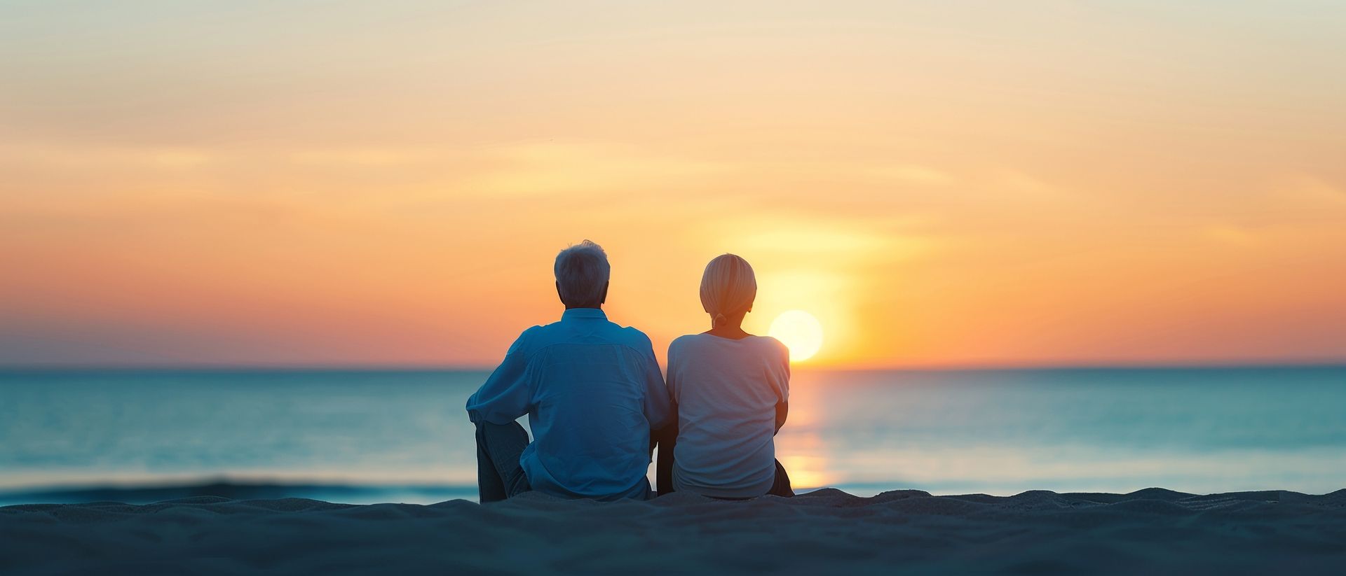 Elderly couple sitting on the beach, facing a vibrant sunset over the ocean.