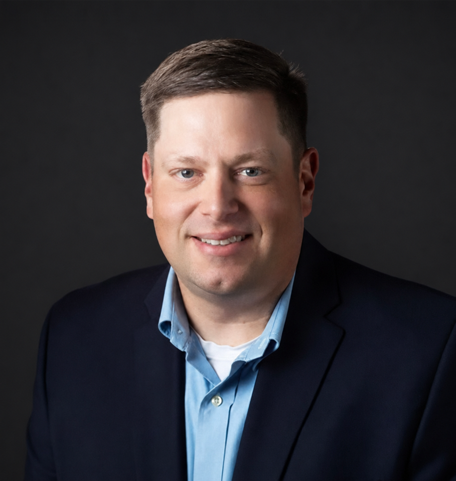 Man in blue shirt and navy blazer smiles against a dark gray background.