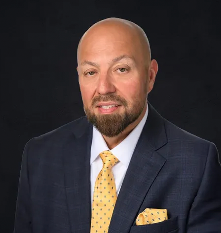 Bald man in navy suit, yellow tie, and beard smiles against a black background.