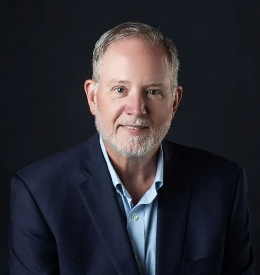 A man with a beard, wearing a blue suit jacket and shirt, smiles against a dark background.