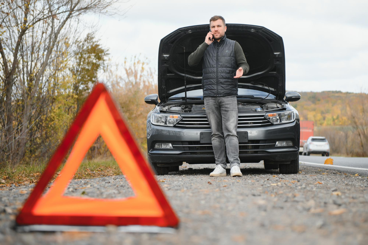 Man with a broken-down car on a roadside calling for help, with a safety triangle in the foreground.