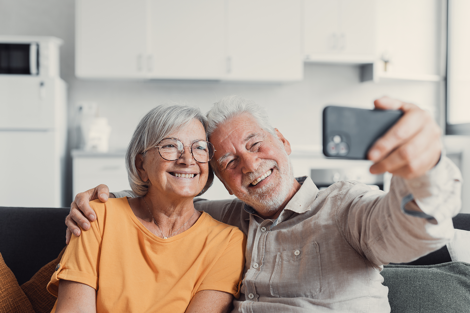 Elderly couple smiling while taking a selfie on a couch in a living room.