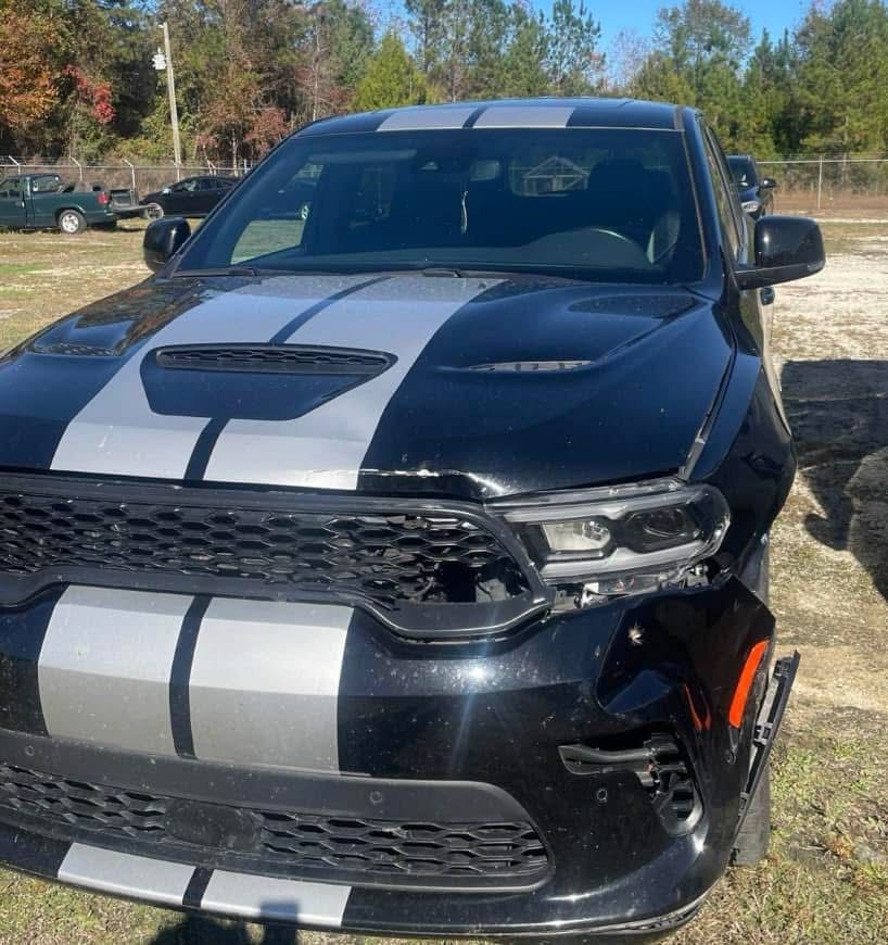 Black and silver SUV with front-end damage; parked on grass near trees, a sunny day.