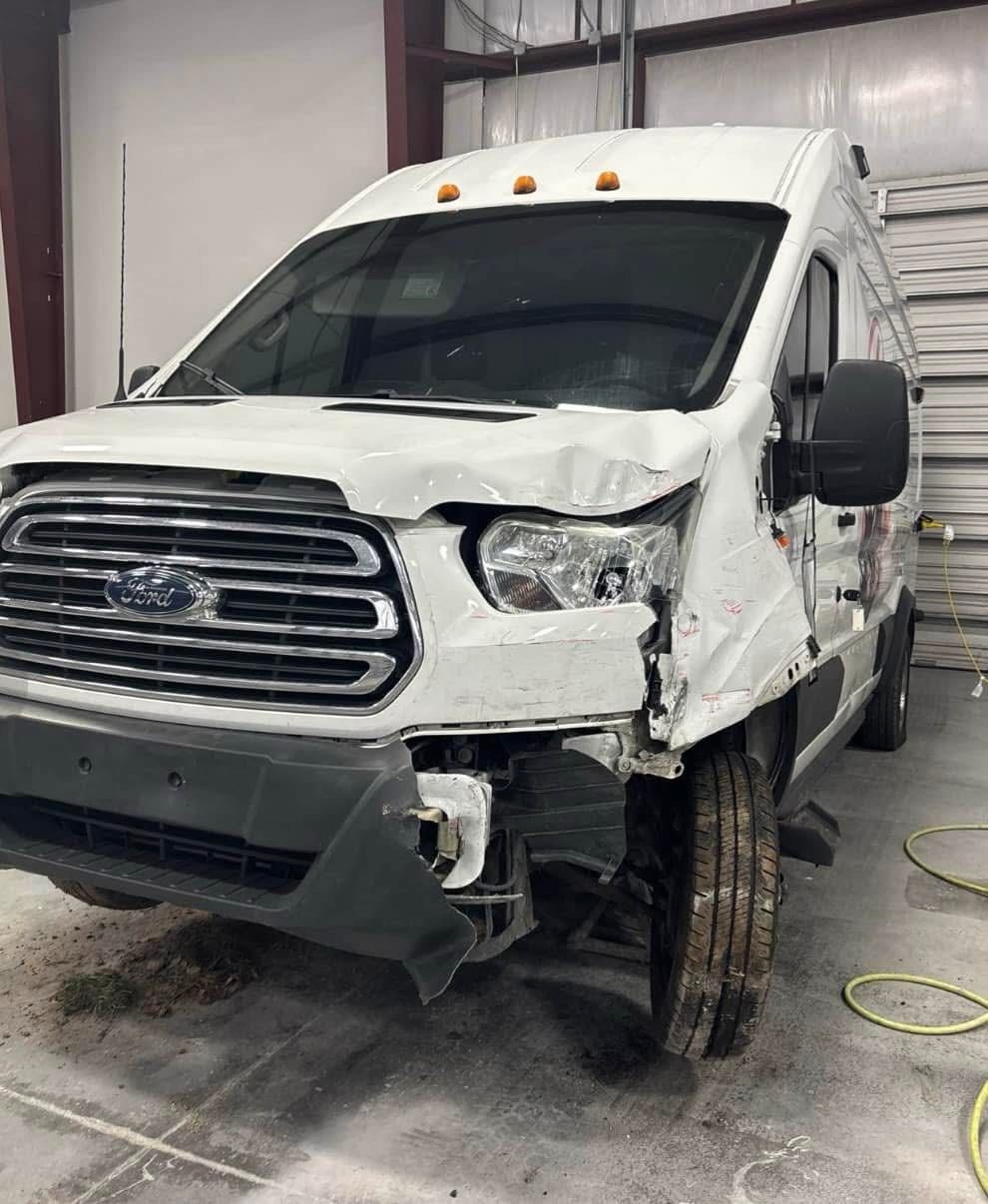 White Ford Transit van with significant front-end damage; damaged headlights, bumper, and fender.