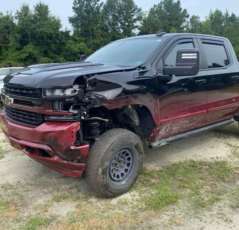 Damaged black and red Chevy pickup truck with front-end collision damage, parked outdoors.