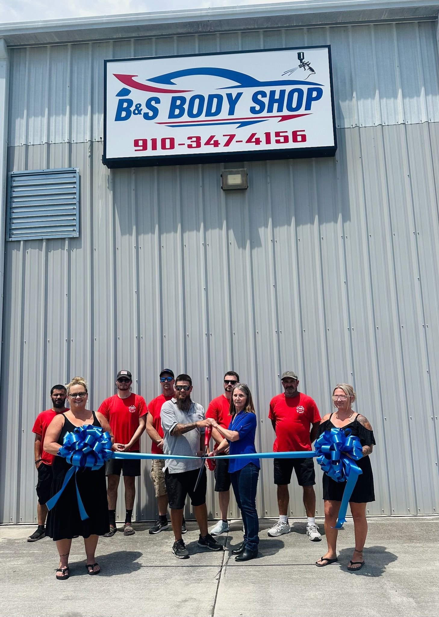 Ribbon-cutting at B&S Body Shop. Group of people in front of a gray building, blue ribbon and bows.