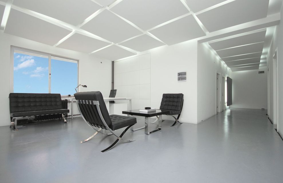 Modern white lounge with black chairs, glass table, large window, and hallway in the background