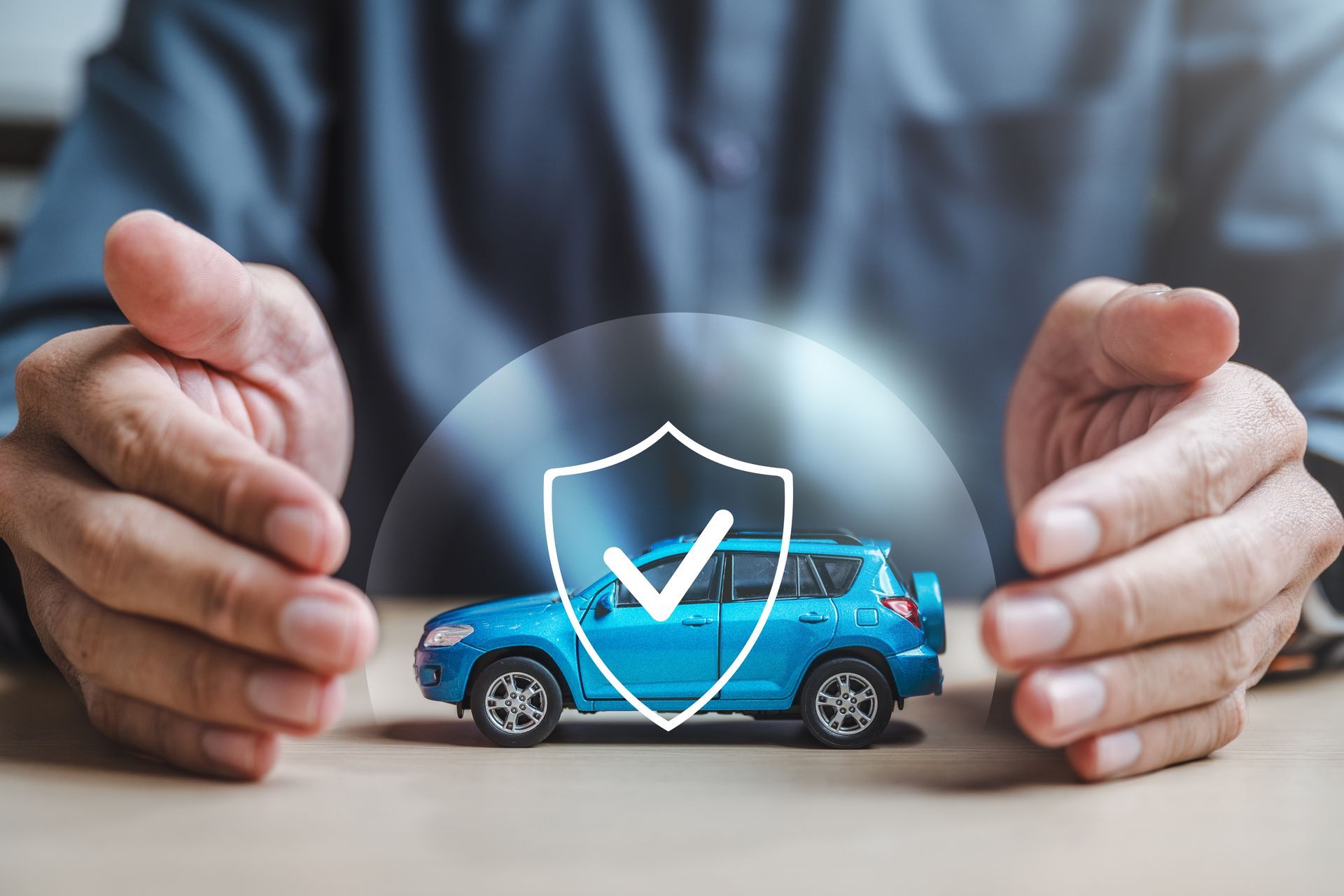 Hands protecting a blue toy car under a shield icon, symbolizing auto insurance or safety
