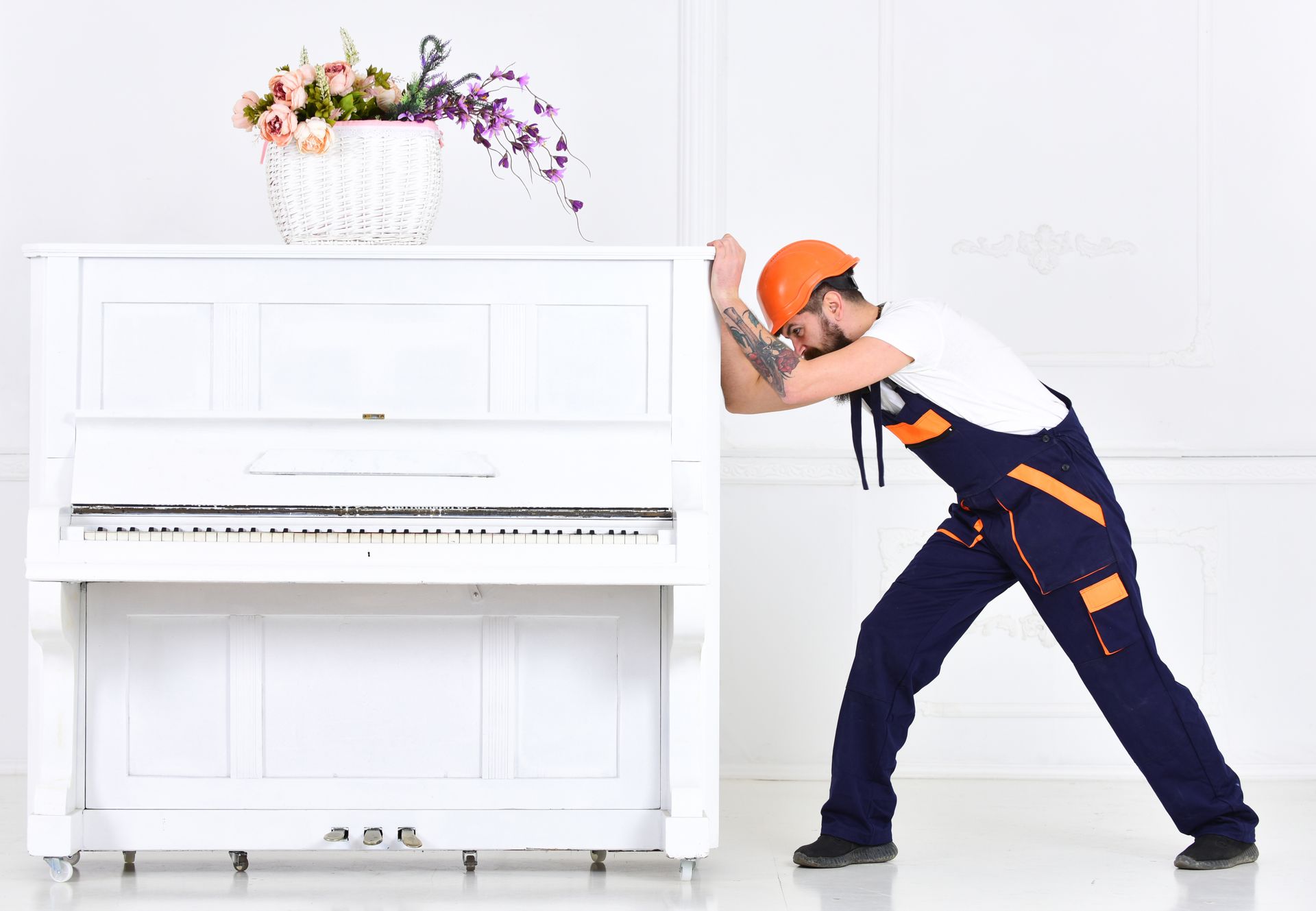One of the piano movers in an orange hard hat pushes a white upright piano with flowers on its lid.