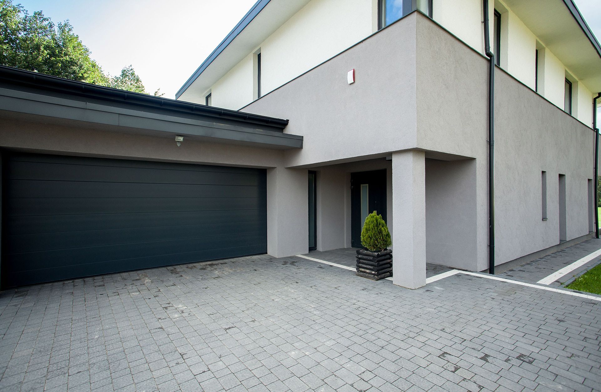 Modern two-story house with a gray garage door, paved driveway, and a small potted tree.