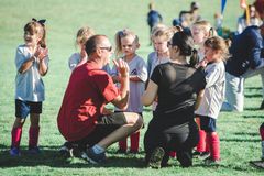 Coach kneels with children on a grassy field during an outdoor sports activity, all gathered in a circle