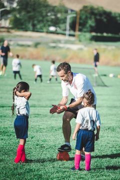 Coach teaching children baseball on a grassy field, with two kids in pink socks listening in front of him