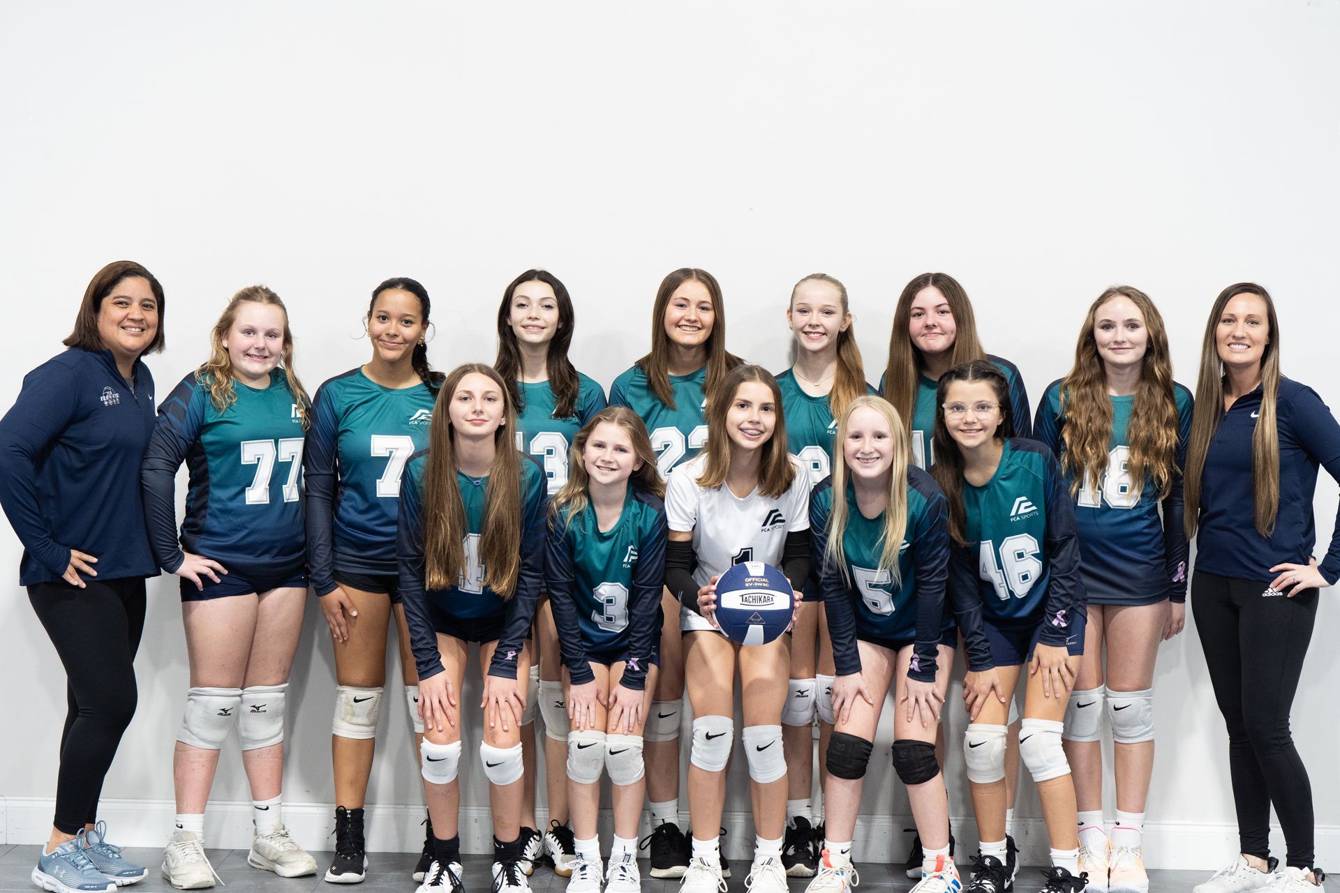 Volleyball team in teal jerseys posing for a group photo against a white backdrop