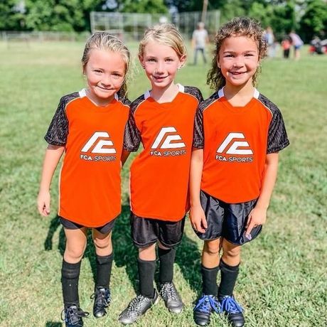 Three youth soccer players in orange uniforms posing on a grassy field