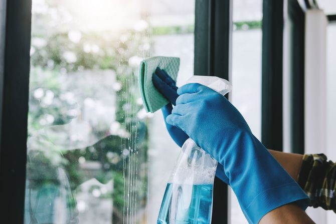 Blue-gloved hand cleaning a window with a spray bottle and squeegee, with bright daylight outside