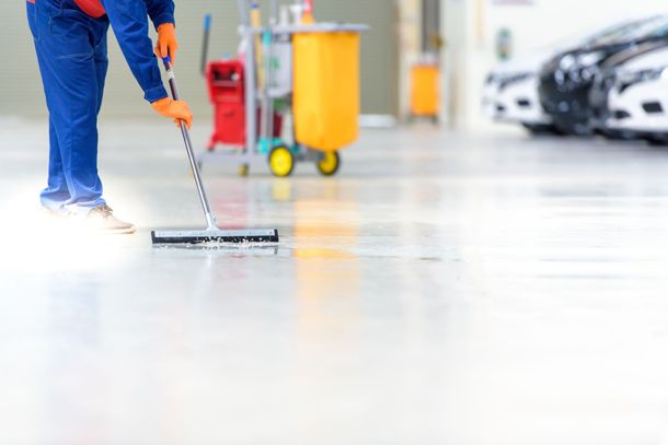 Worker cleaning a bright garage floor with a mop beside parked cars and a yellow trash bin