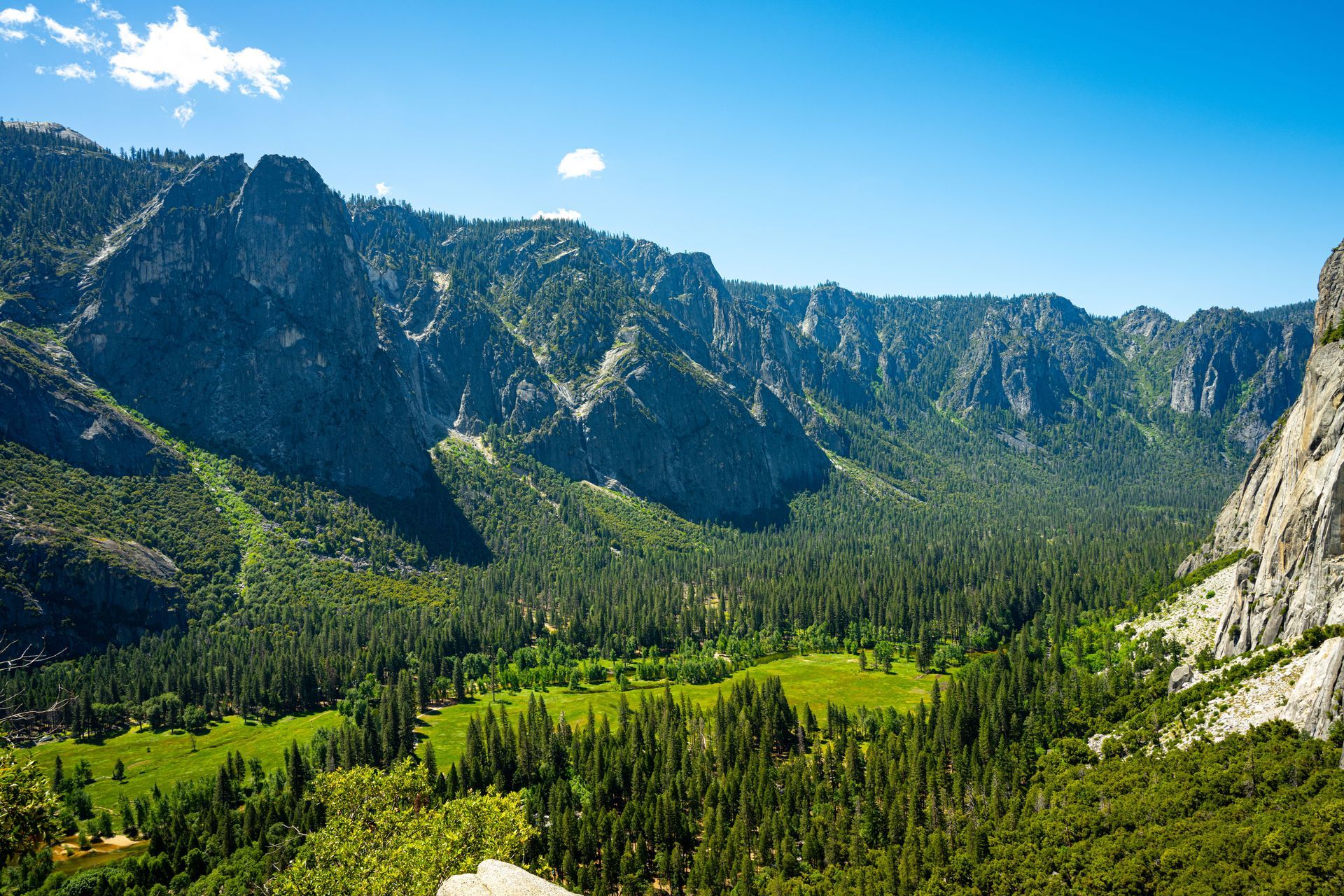 Mountain valley with green forest, rocky cliffs, and bright blue sky.