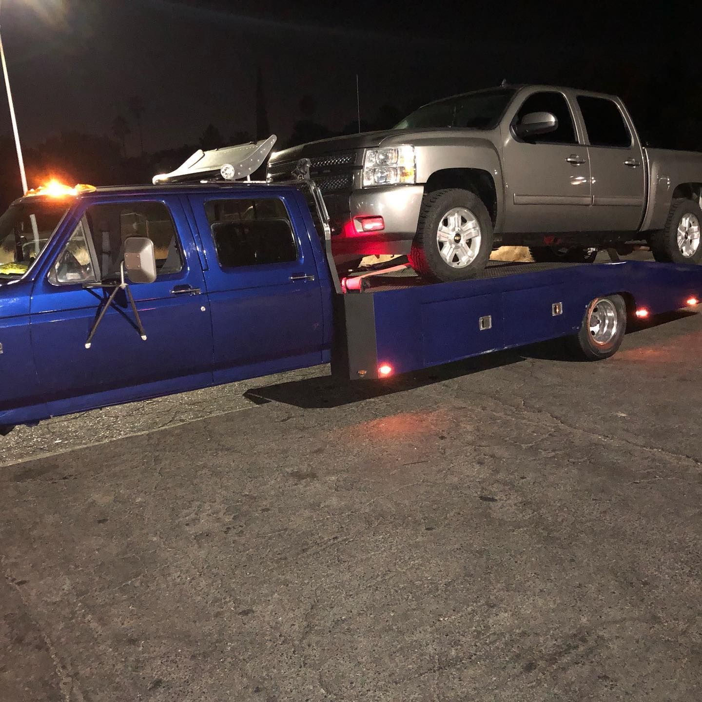 Blue tow truck carrying a silver pickup on a flatbed at night in a parking lot
