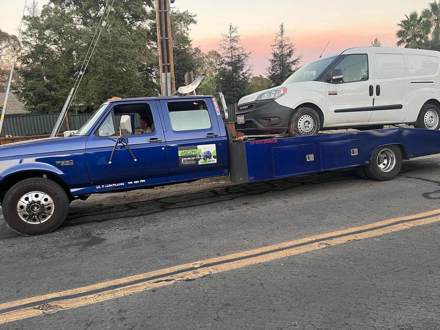 Blue tow truck carrying a white van on a flatbed at dusk by a road
