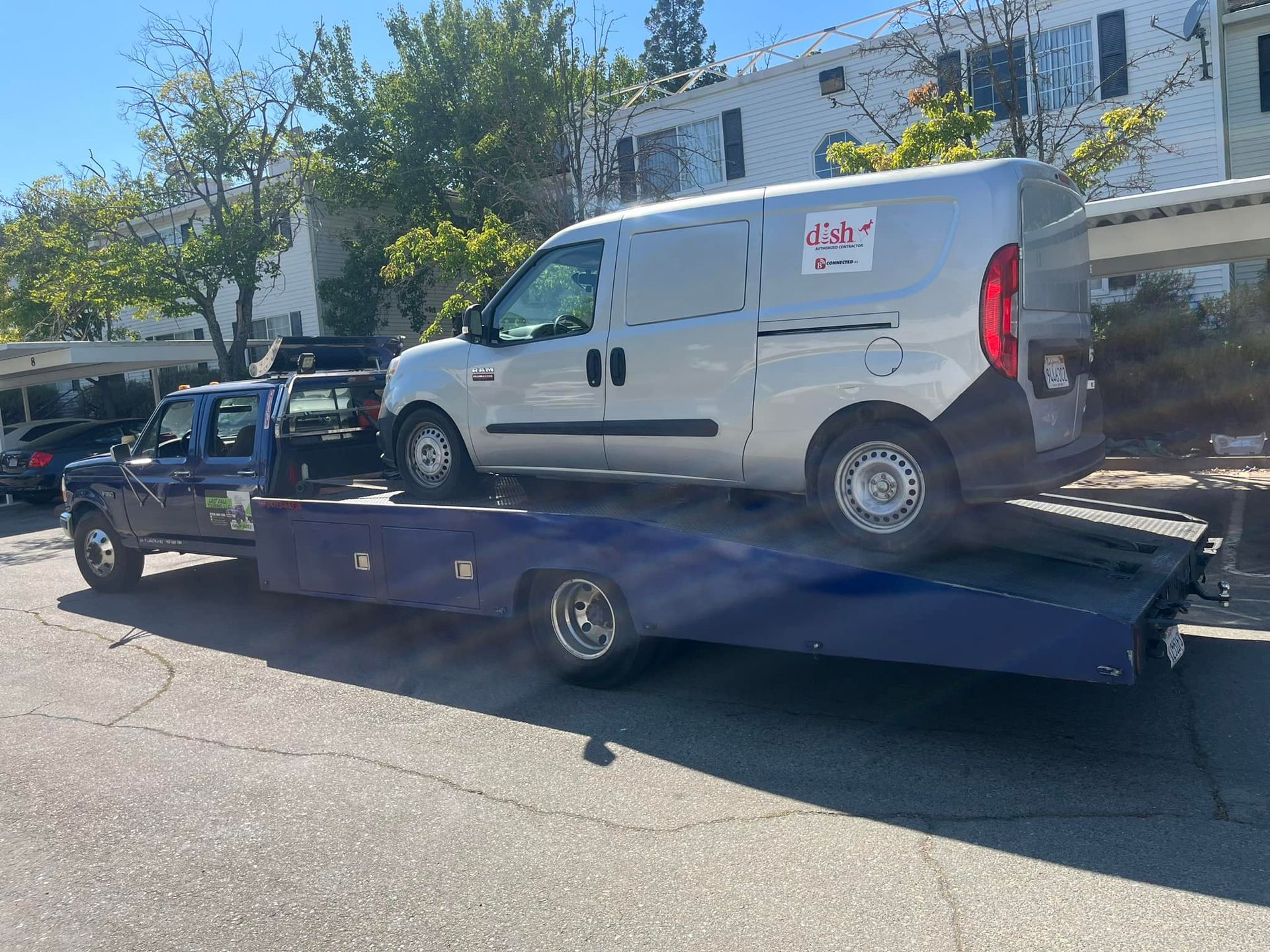 White van loaded on a blue flatbed tow truck on a sunny street beside buildings and trees