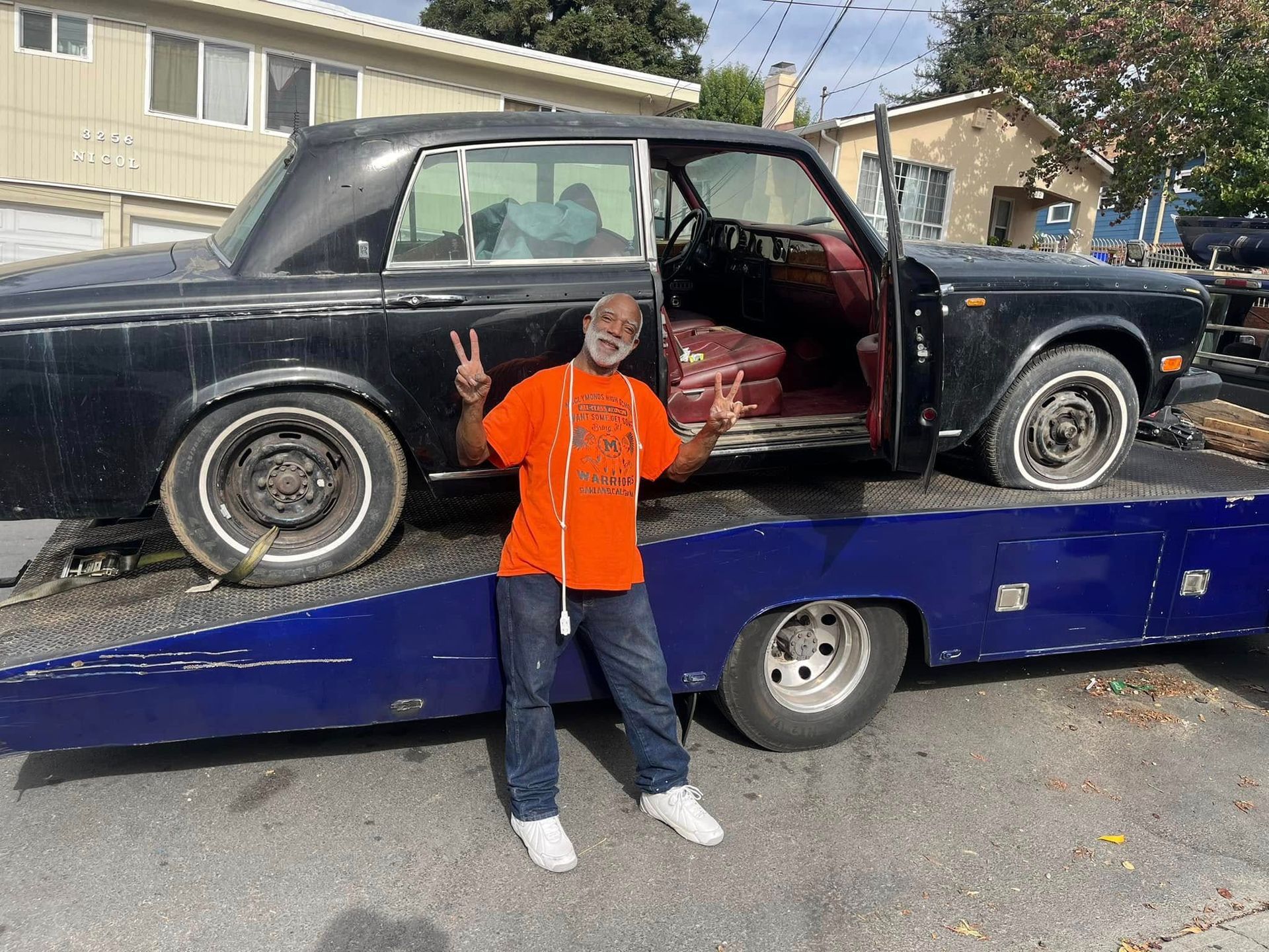 Person in orange jacket posing beside a black classic car on a blue trailer in a driveway.