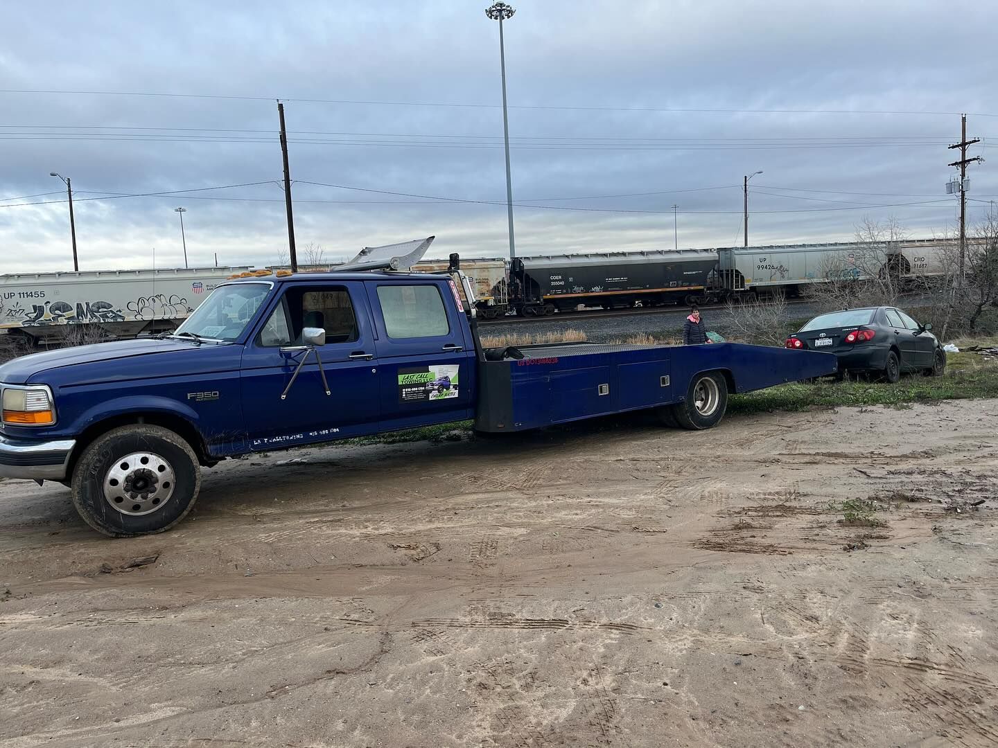 Blue tow truck hauling a black sedan on a flatbed beside a rocky roadside under a cloudy sky
