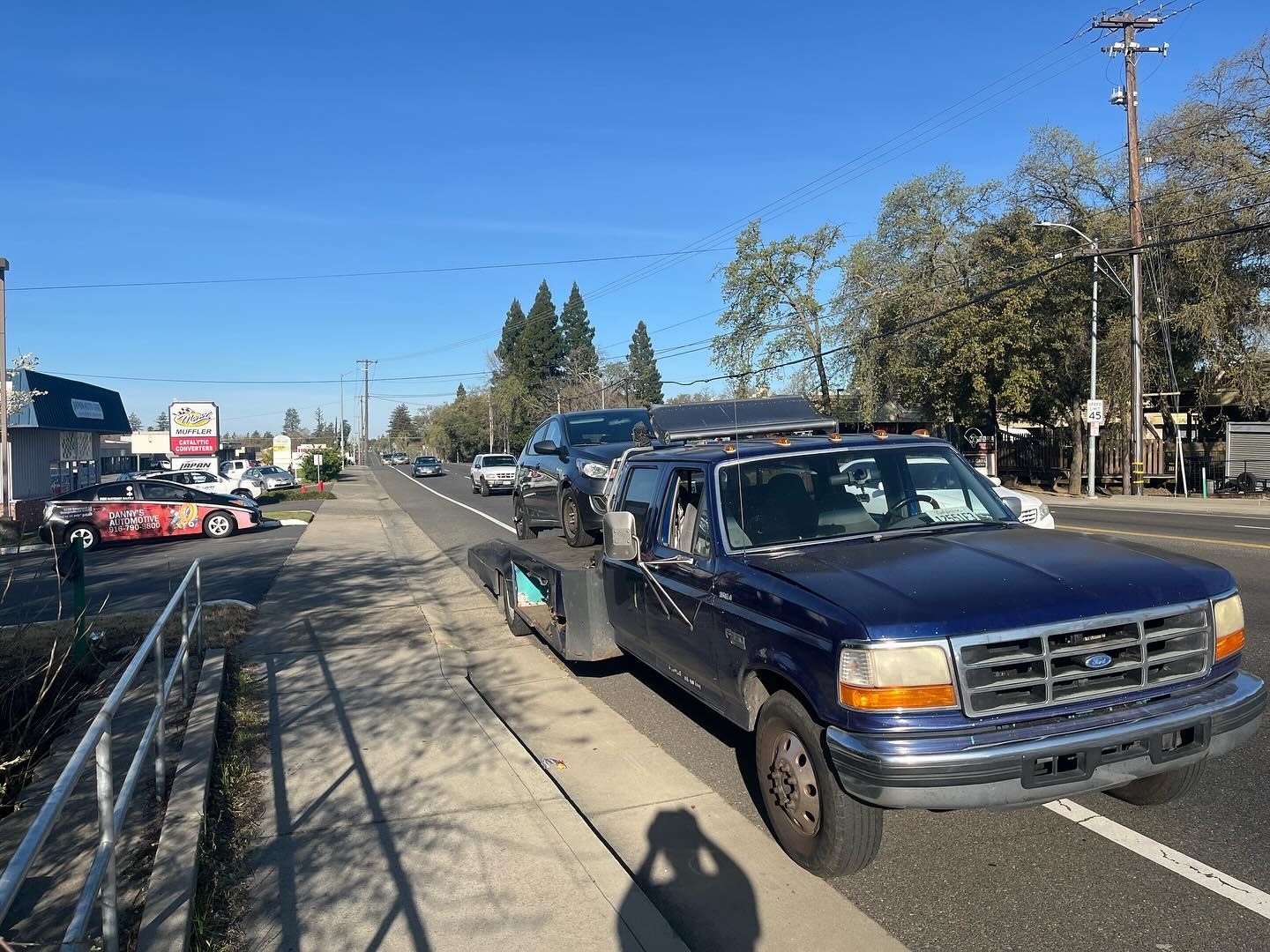 Blue tow truck parked on a sunny roadside with cars, trees, and utility poles along the street.