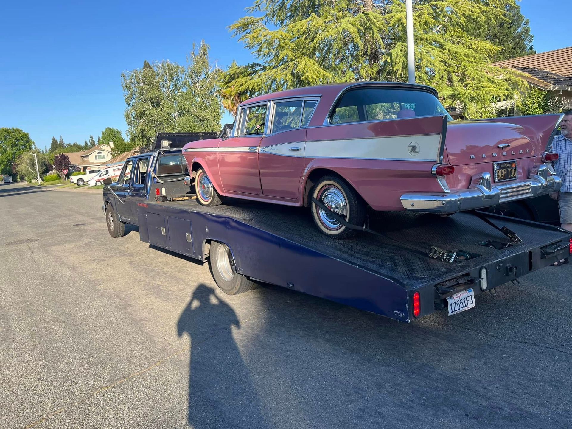 Pink-and-white classic car on a black trailer parked on a residential street, with trees and houses nearby