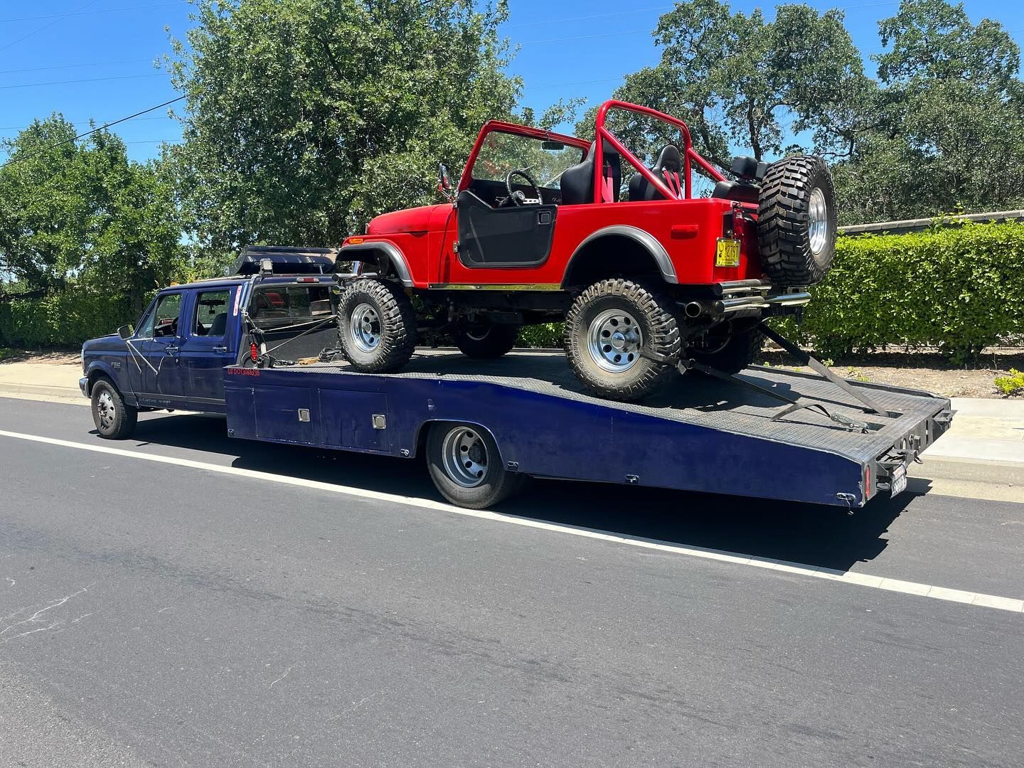 Red off-road Jeep strapped on a blue tow truck driving on a sunny road.