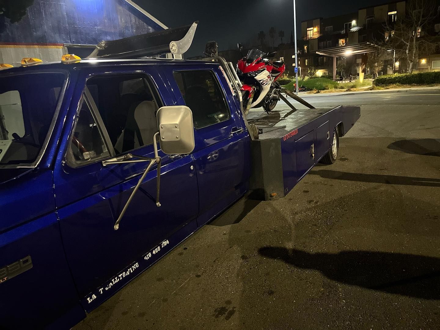 Blue pickup truck with a crumpled side and a motorcycle loaded on the back at night in a parking lot