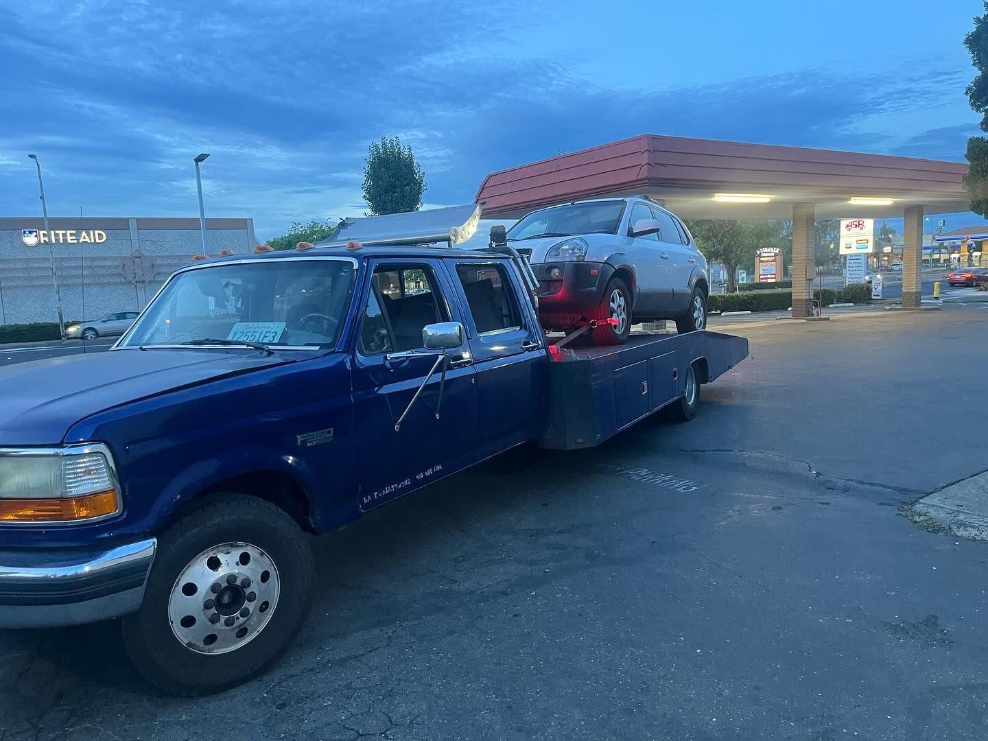 Blue tow truck carrying a car on a flatbed at a gas station at dusk